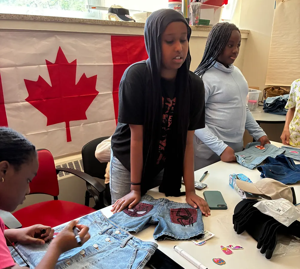Three youth at a table, one sitting, modifying jean shorts with their own designs