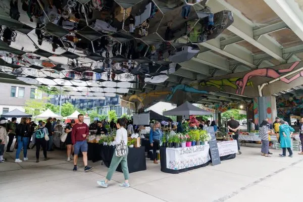 farmers' market under expressway, people shopping, booths in a vibrant scene with murals and art.