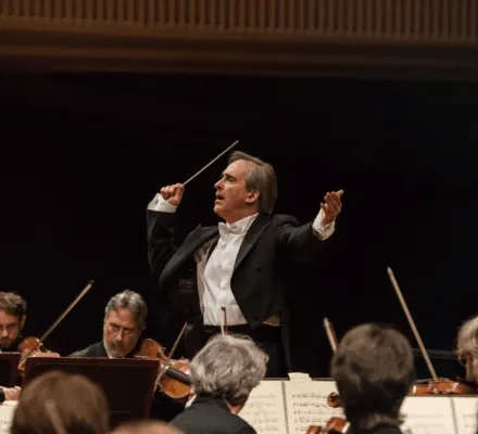James Conlon performs on stage. He wears a full suit, with his hand raised with a baton.