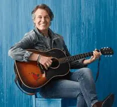 Jim Cuddy holds a guitar in his lap. He wears a jean jacket and jeans and sits in front of a blue background.