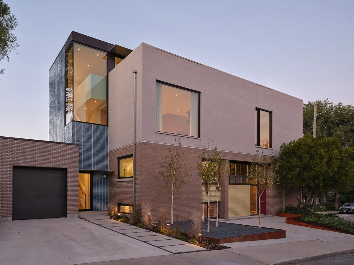 Modern two-storey brick and stucco house at dusk, with illuminated glass stairwell tower and side garage.