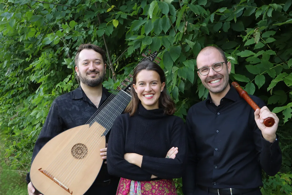 Three smiling musicians pose against green foliage: man holding lute, woman in center and man holding wooden recorder.
