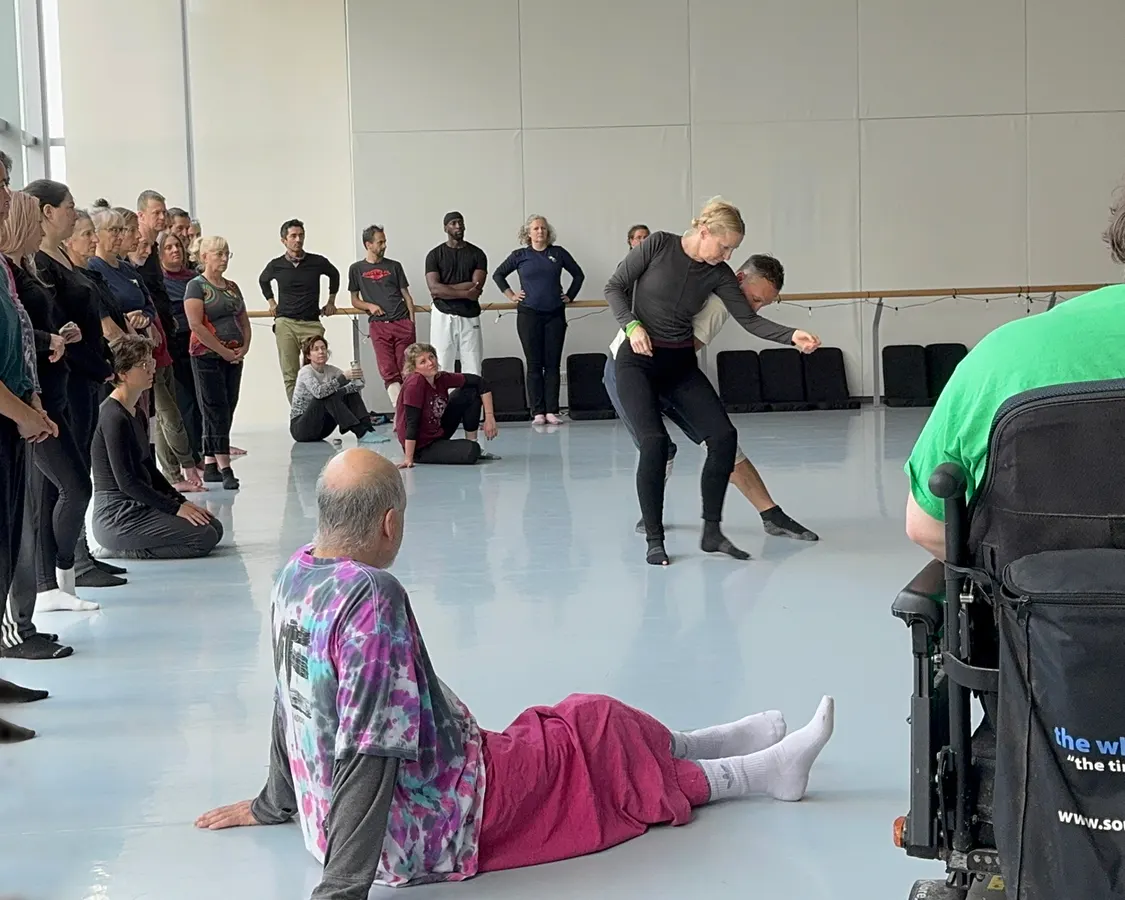A dance group stands near the barre, watching an instructor demonstrate moves.