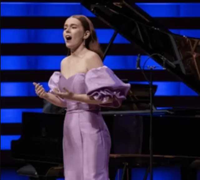 A woman wearing a lilac colour evening dress sings in front of a grand piano.