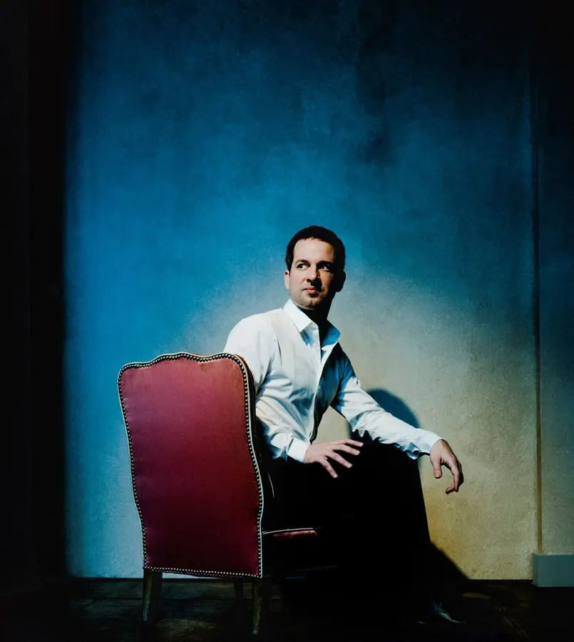 Bertrand Chamayou sits on a red chair with a spotlight shinning at him over a blue background